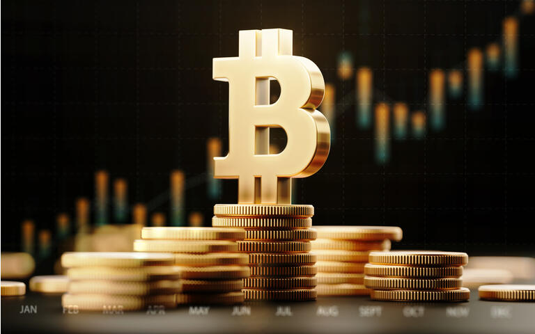 Picture of a bitcoin symbol standing on top of stacked gold coins, with a red candlestick chart behind it